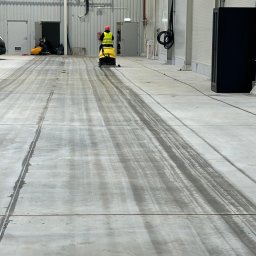 SOLIDTRANS-TEAM ILONA ARCISZEWSKA - Pracownik w kamizelce odblaskowej czyści posadzkę przemysłową za pomocą maszyny czyszczącej. Widoczne ślady po czyszczeniu, hala z metalowymi ścianami.