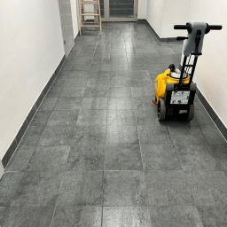 SOLIDTRANS-TEAM ILONA ARCISZEWSKA - Wypolerowana, szara podłoga z płytek w korytarzu biurowym po czyszczeniu maszynowym. W tle drabina i przeszklone drzwi.