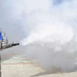 Osoba w kombinezonie ochronnym rozpyla gęstą mgłę z przenośnego zamgławiacza na zewnątrz.