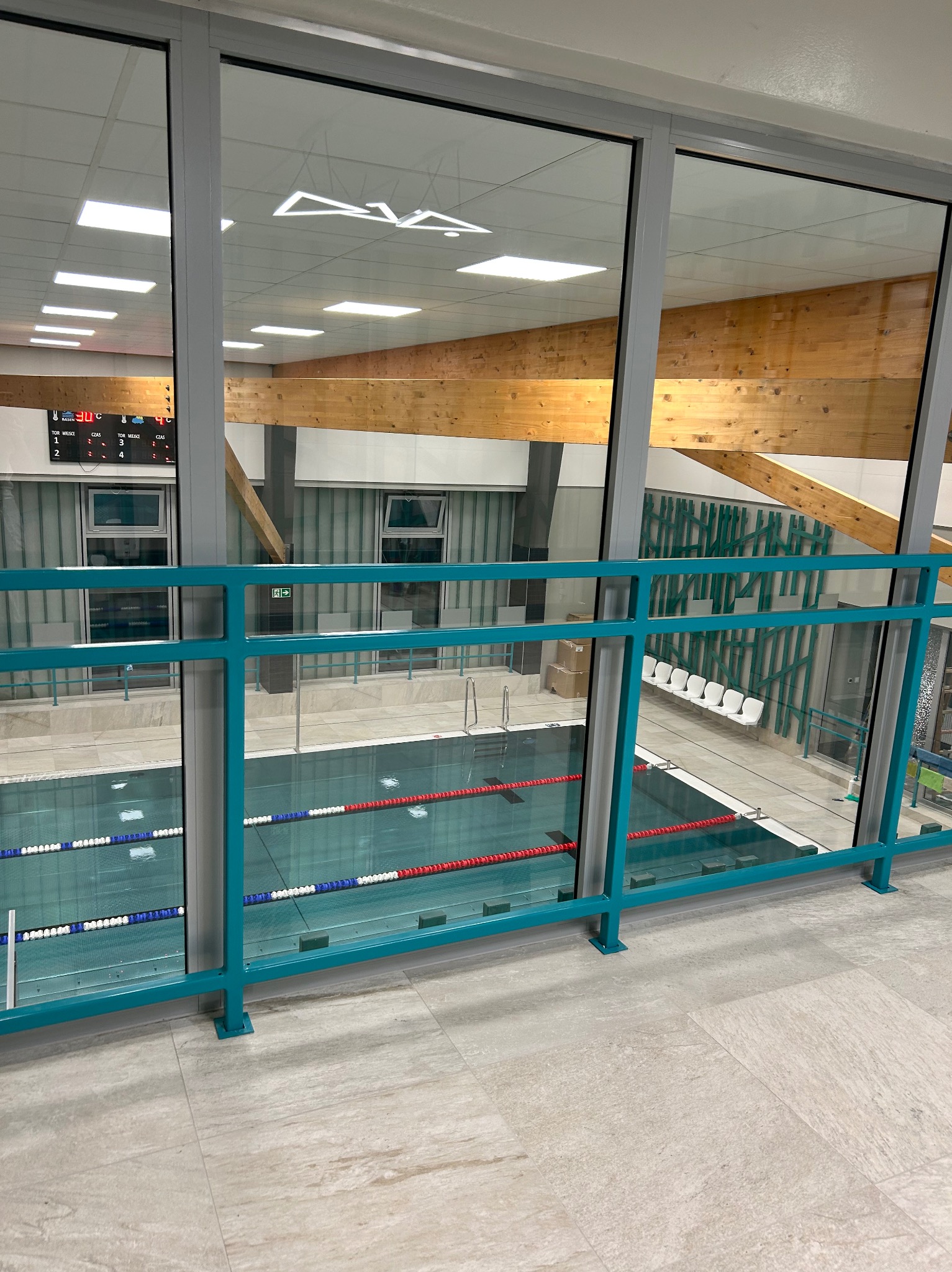Widok na basen sportowy z antresoli przez szklaną balustradę. Widoczne tory pływackie, tablica wyników i drewniane elementy konstrukcyjne dachu.