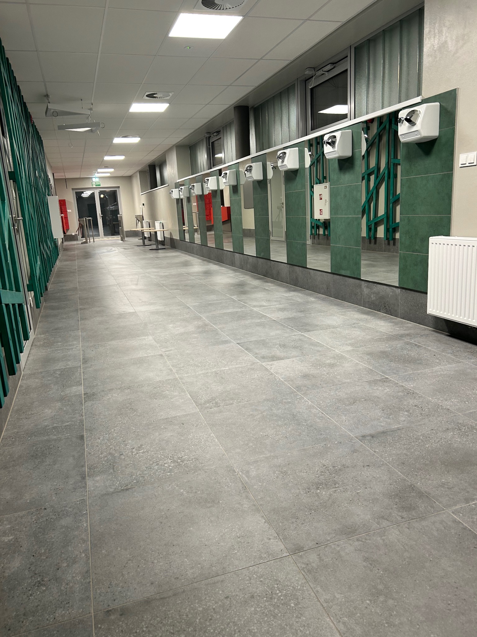 Wnętrze publicznej toalety z rzędem umywalek i suszarek do rąk, szarą podłogą z płytek i zielonymi akcentami. Widok na korytarz z wyjściem ewakuacyjnym.