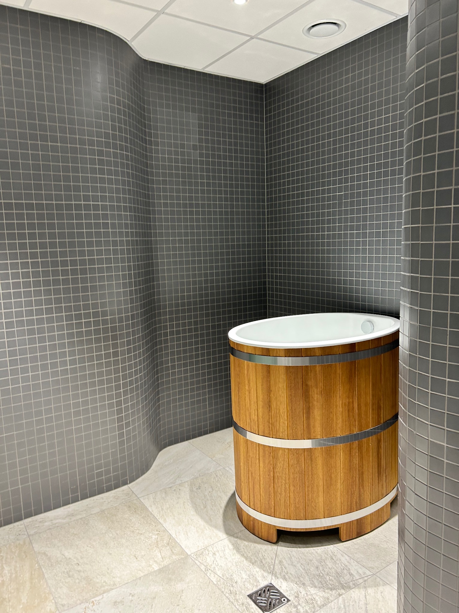 Drewniana balia z metalowymi obręczami w nowoczesnej łazience z szarymi, kwadratowymi płytkami i beżową podłogą. Widoczny odpływ podłogowy.