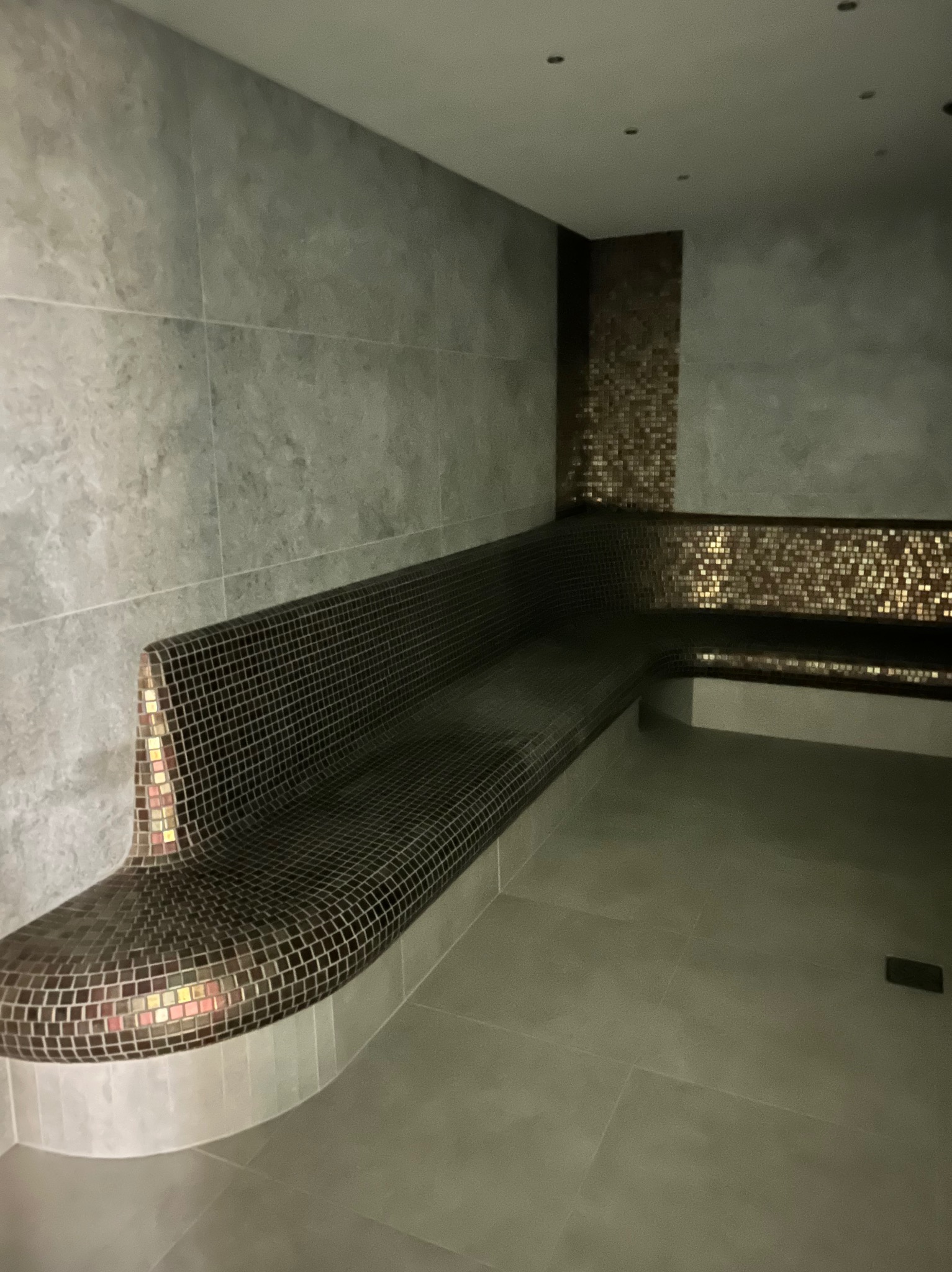 Mozaika w strefie relaksu: wykończone płytkami siedziska i ściany w odcieniach szarości i brązu, tworzące spójną, elegancką przestrzeń. Widok z perspektywy.