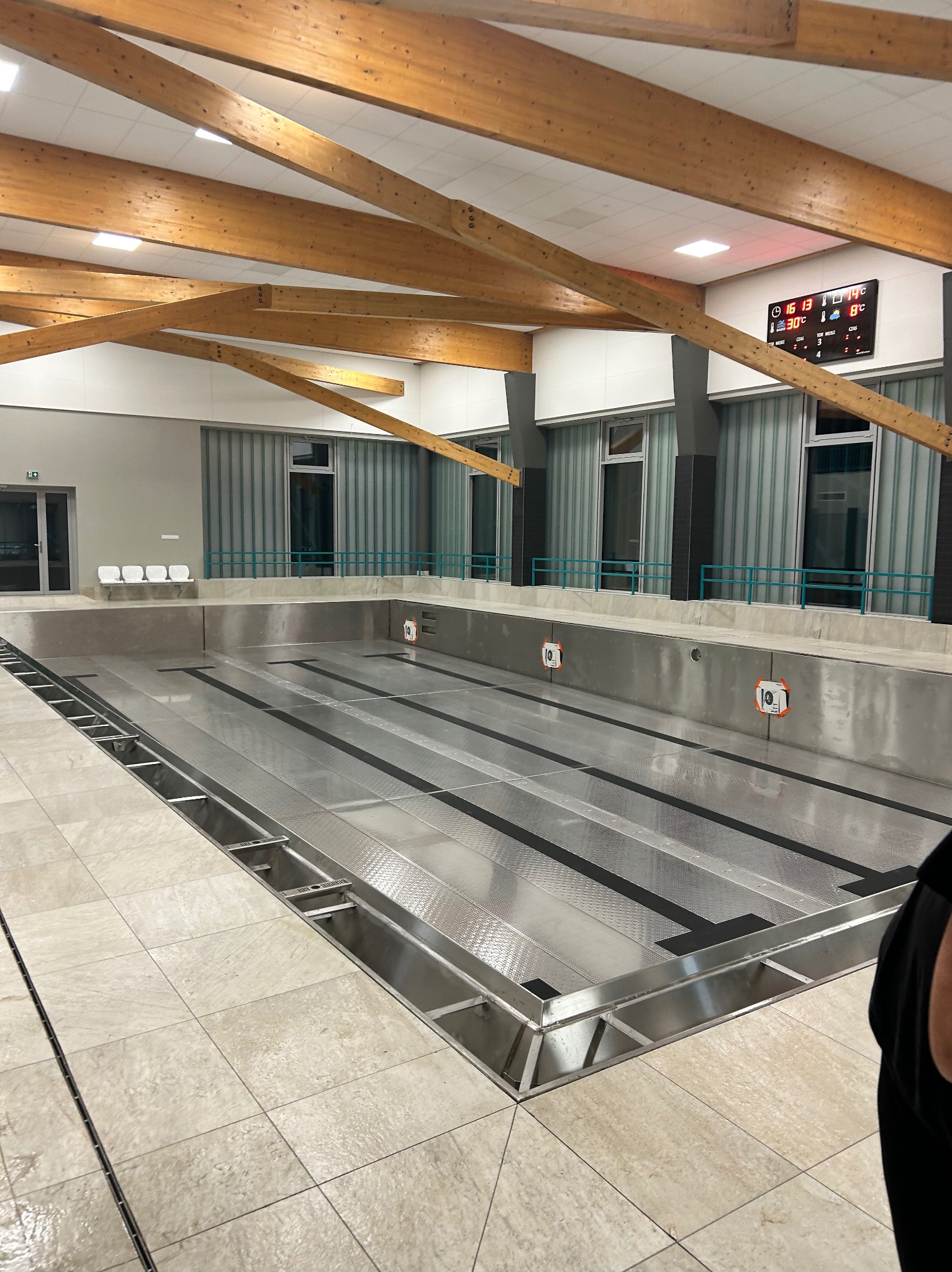 Opróżniony basen sportowy z metalowym dnem i drewnianą konstrukcją dachu. Widoczne okna, płytki podłogowe i tablica wyników.