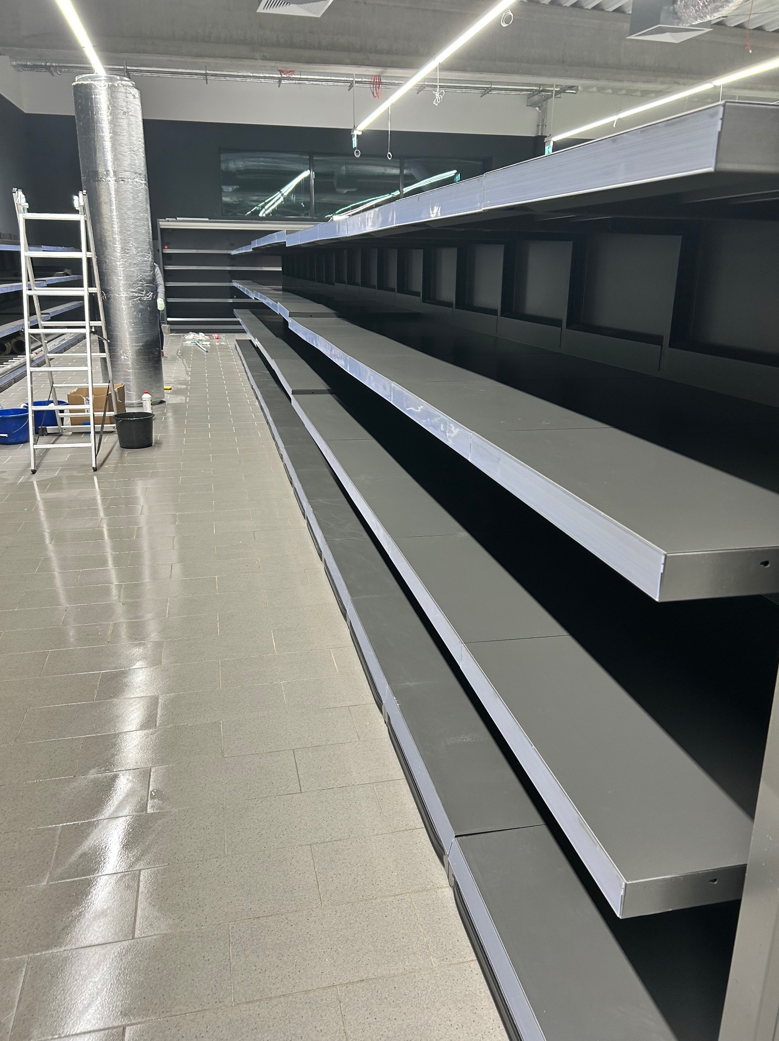 Puste, szare półki w sklepie po remoncie, widoczna drabina i zabezpieczona kolumna. Podłoga umyta, brak towaru. Przygotowanie do otwarcia sklepu.