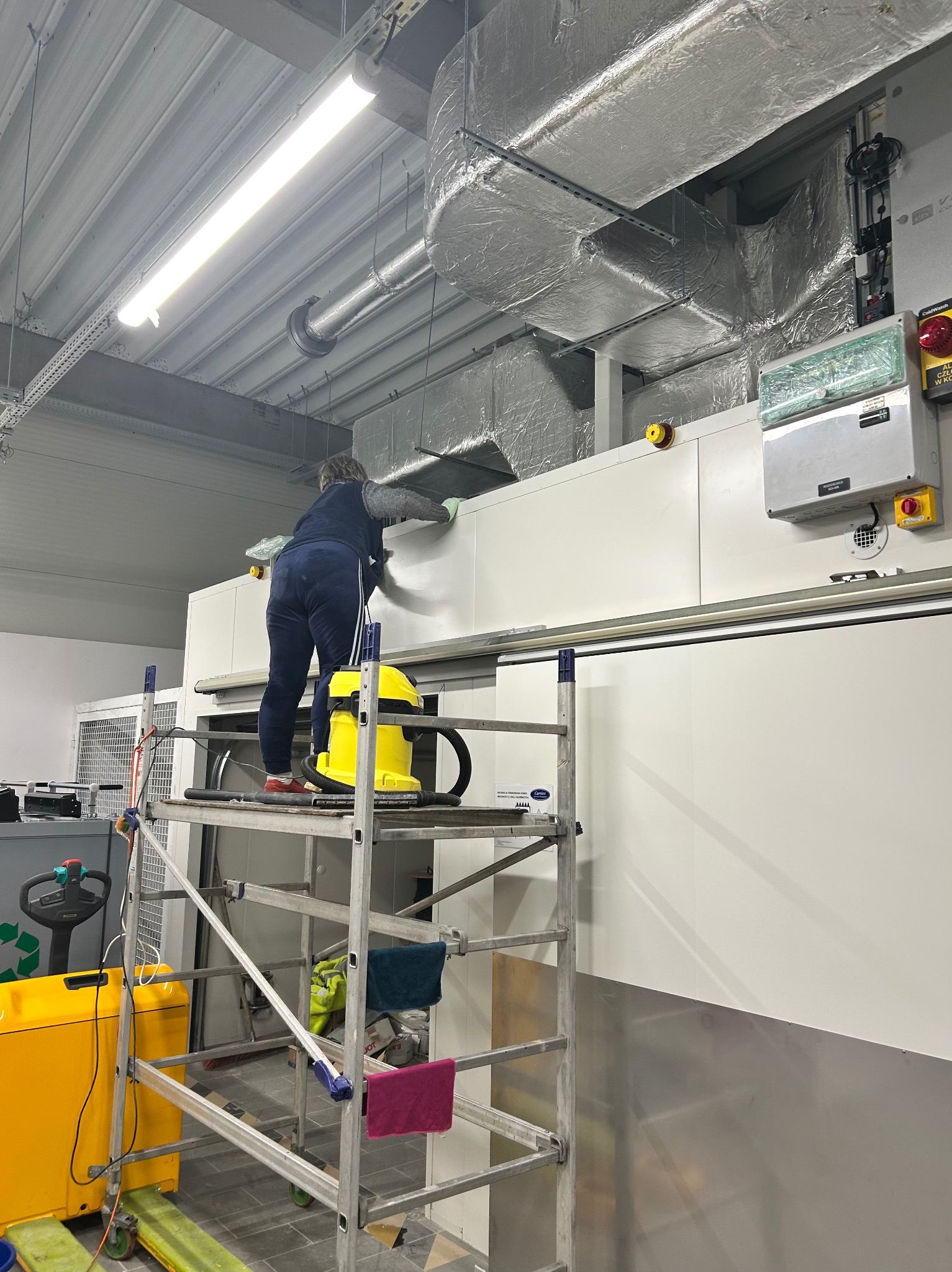 Pracownik na rusztowaniu czyści białe panele w hali przemysłowej. Widoczny żółty odkurzacz przemysłowy i elementy wentylacji pod sufitem.