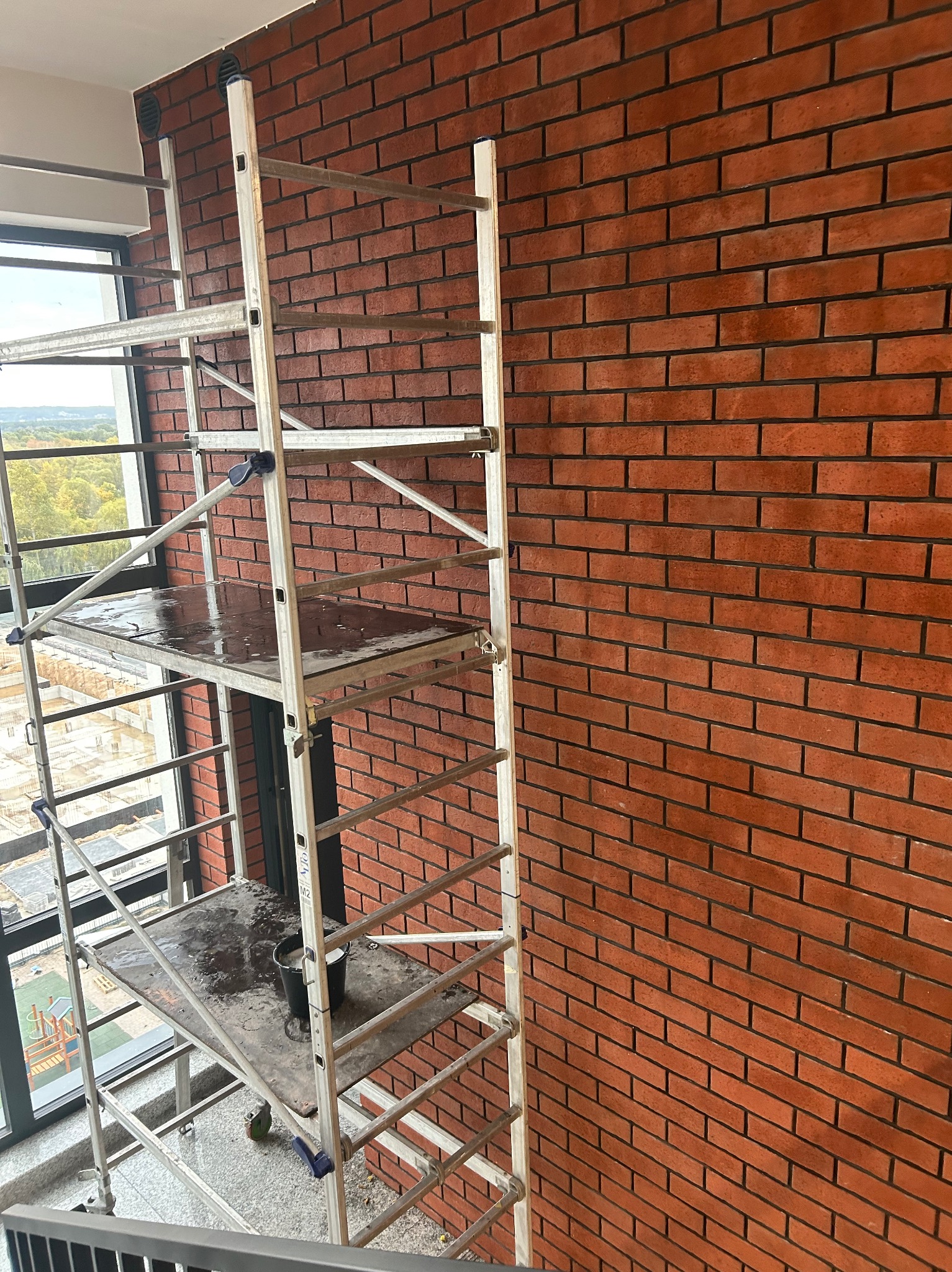 Ceglana ściana po czyszczeniu, widoczna z rusztowania. Na podeście wiadro z wodą. Okno z widokiem na zewnątrz. Detal po zakończonych pracach.
