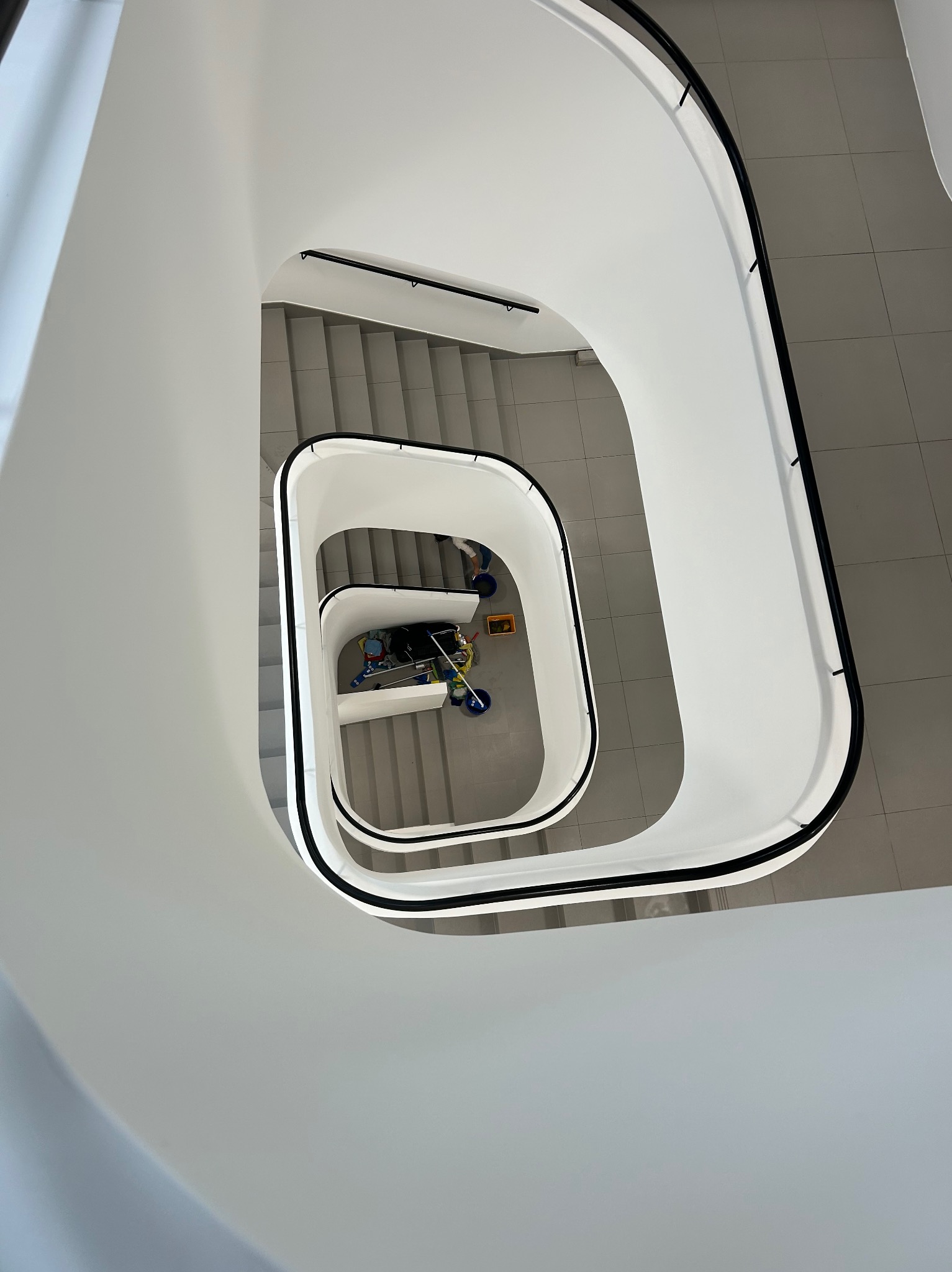 Spiralne schody z widokiem z góry na wózek do sprzątania na podeście. Minimalistyczna architektura wnętrza w jasnych barwach.