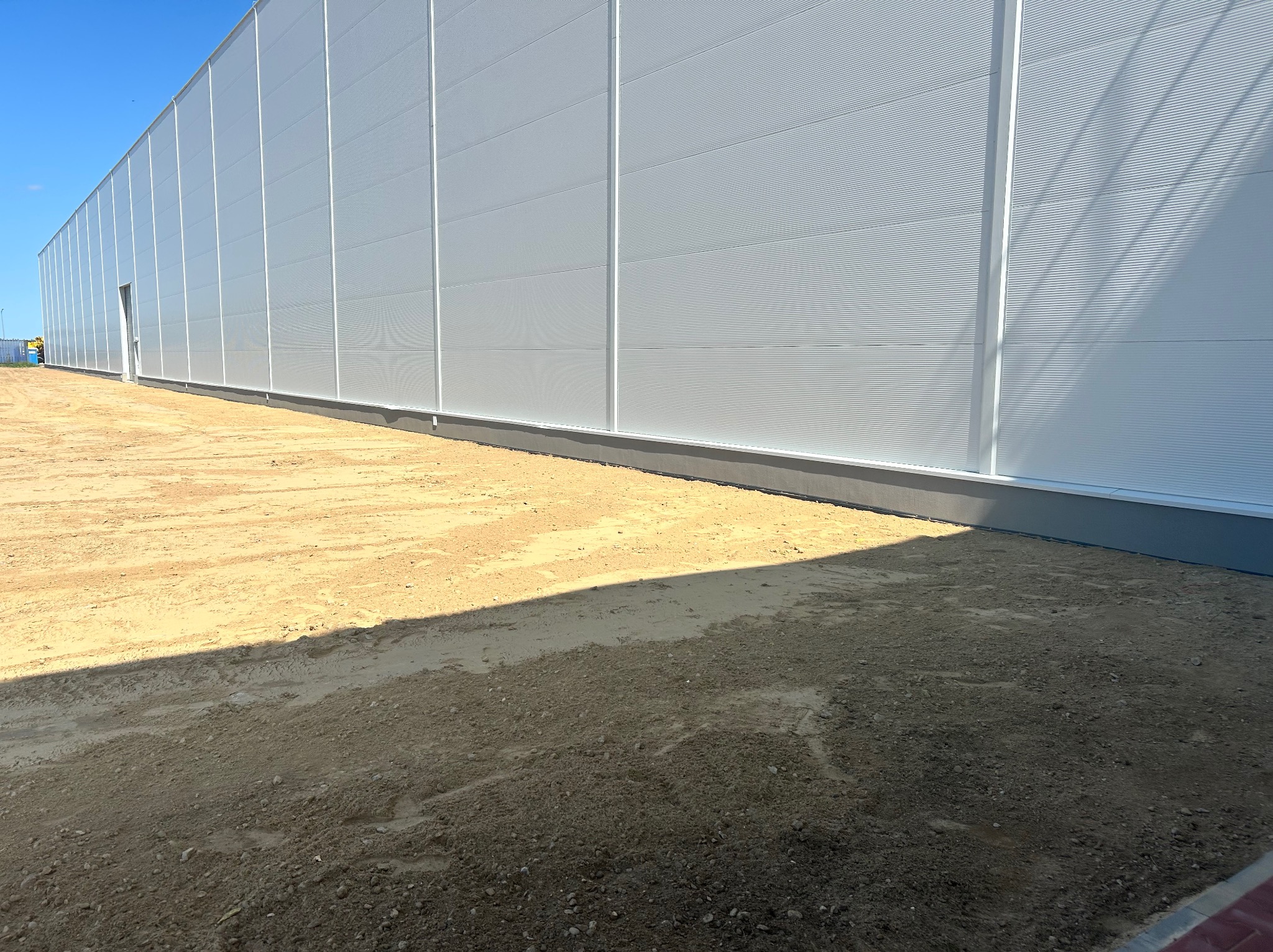 Biała hala magazynowa z panelami, widok z zewnątrz, teren przed budynkiem z piaskiem i cieniem. Minimalistyczna architektura przemysłowa w słoneczny dzień.