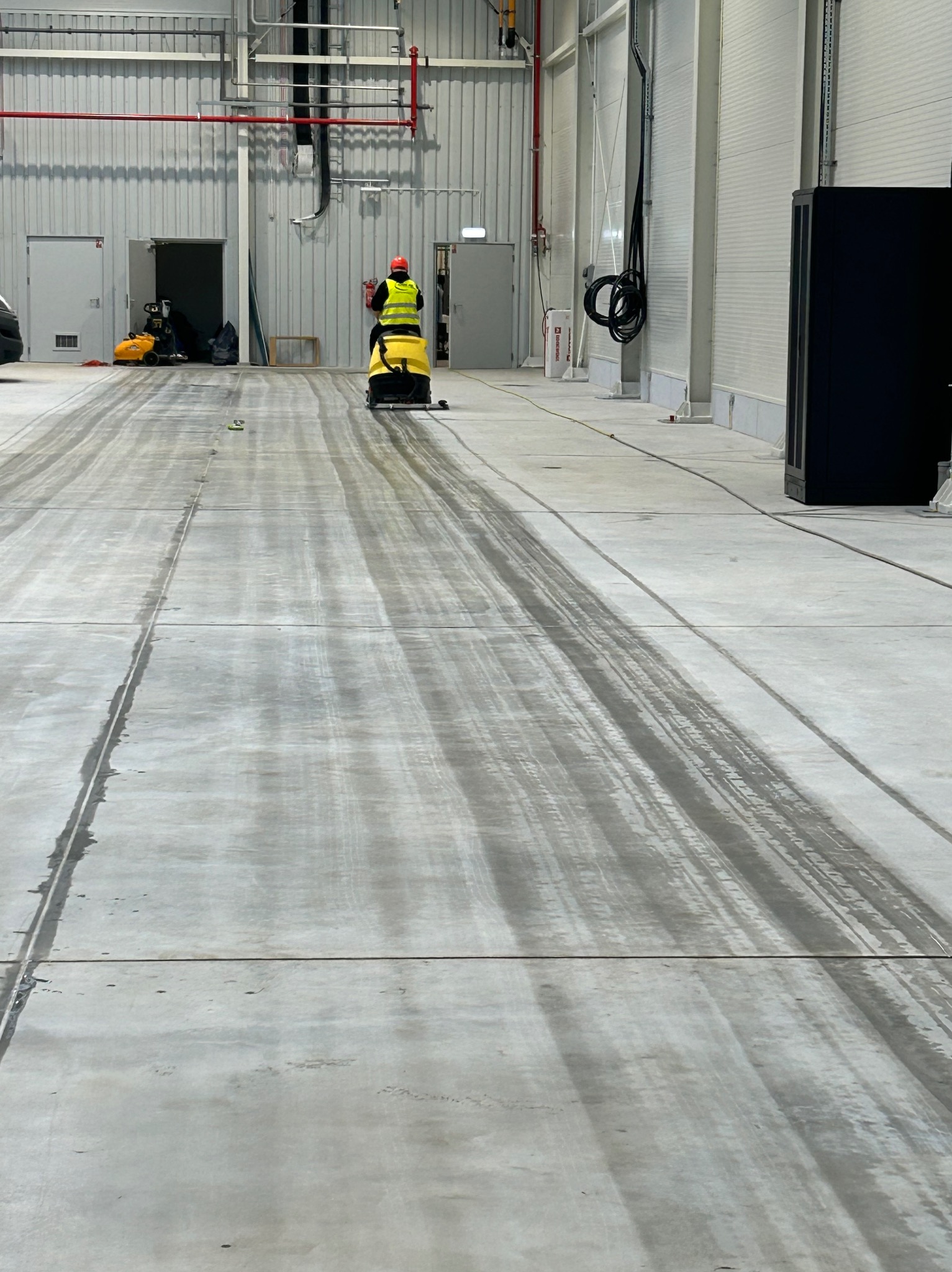 Pracownik w kamizelce odblaskowej czyści posadzkę przemysłową za pomocą maszyny czyszczącej. Widoczne ślady po czyszczeniu, hala z metalowymi ścianami.