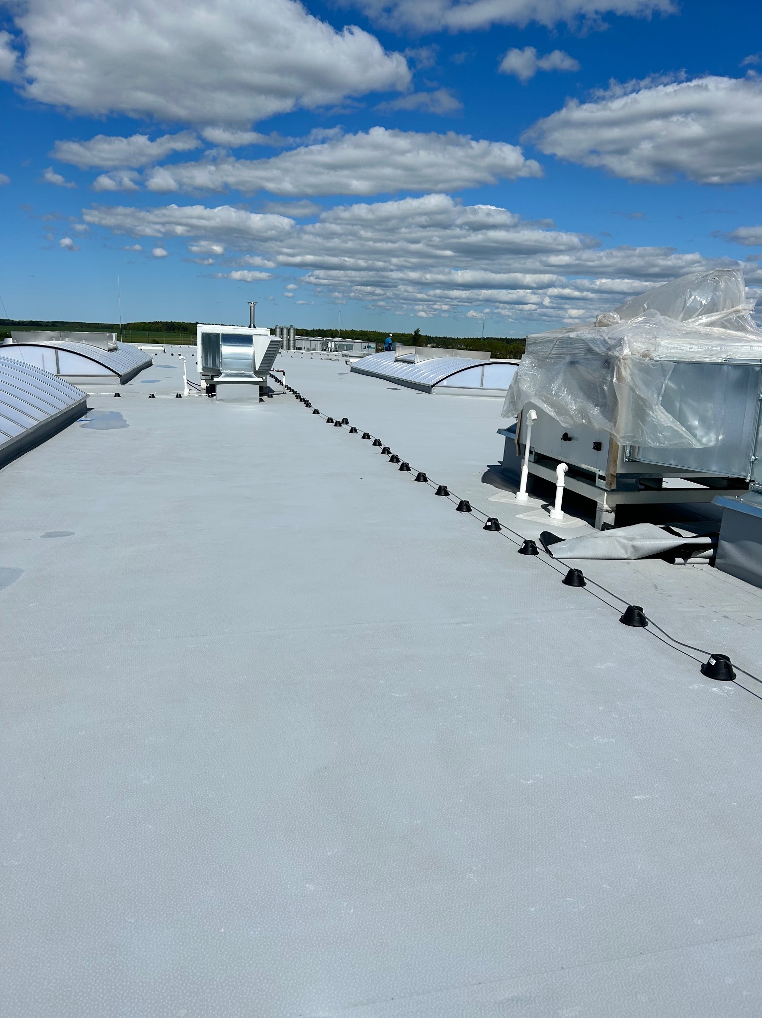 Szary dach przemysłowy z elementami wentylacji i zabezpieczeniami odgromowymi na tle błękitnego nieba z chmurami. Widok z góry, szeroki kadr.