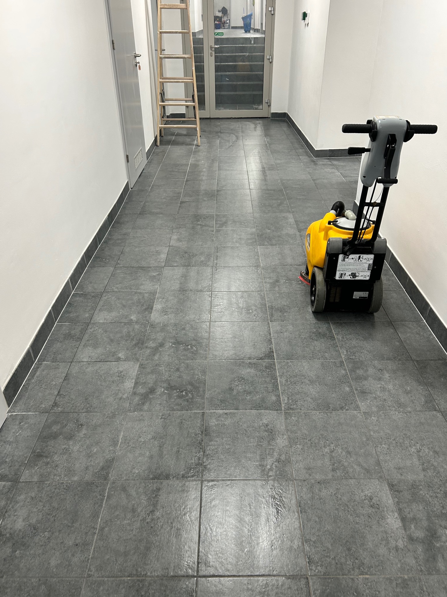 Wypolerowana, szara podłoga z płytek w korytarzu biurowym po czyszczeniu maszynowym. W tle drabina i przeszklone drzwi.