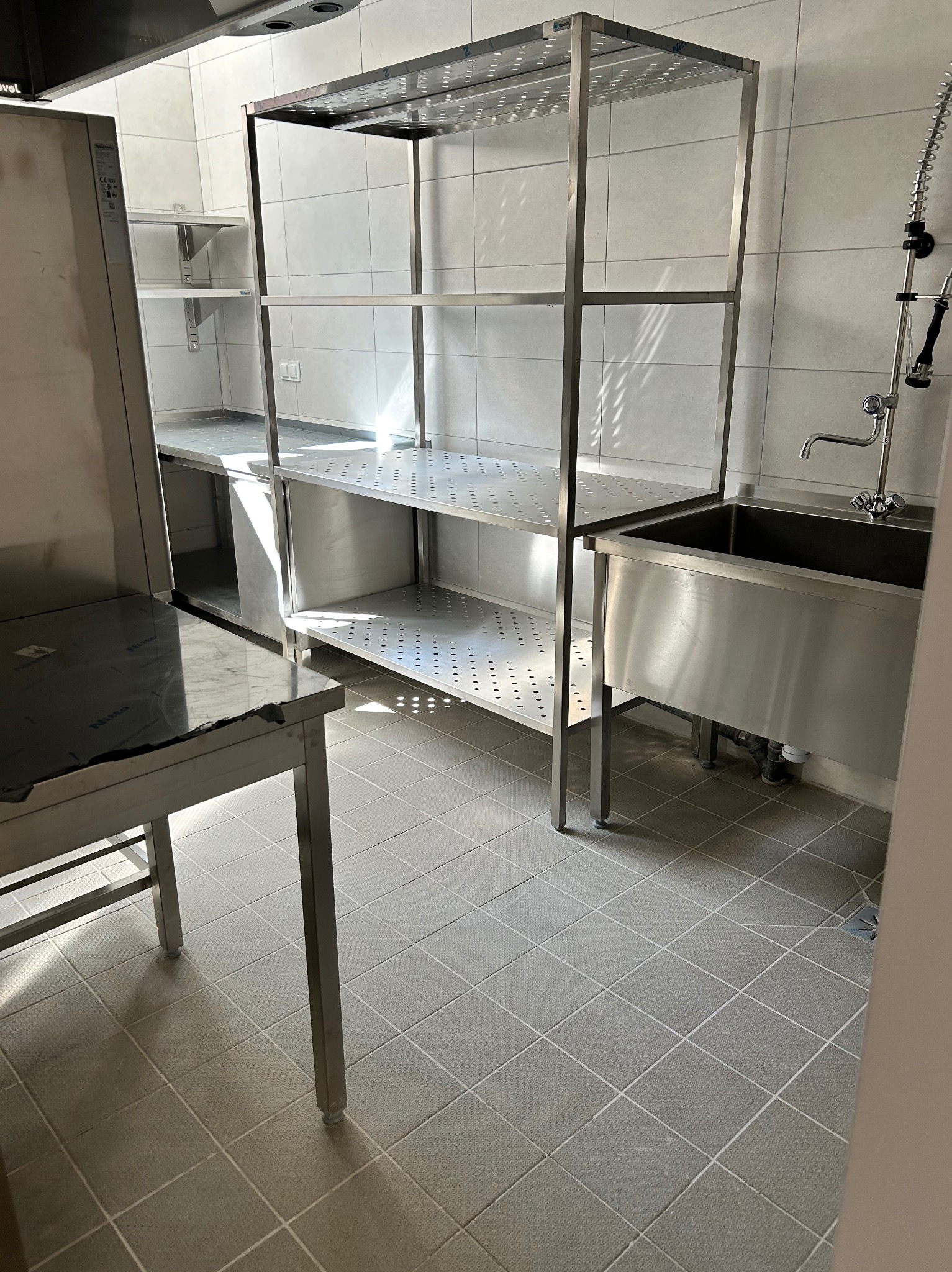 Wnętrze kuchni po sprzątaniu: błyszczące, metalowe regały, zlew i stół na tle jasnych, wykafelkowanych ścian i podłogi. Widoczne detale wyposażenia.
