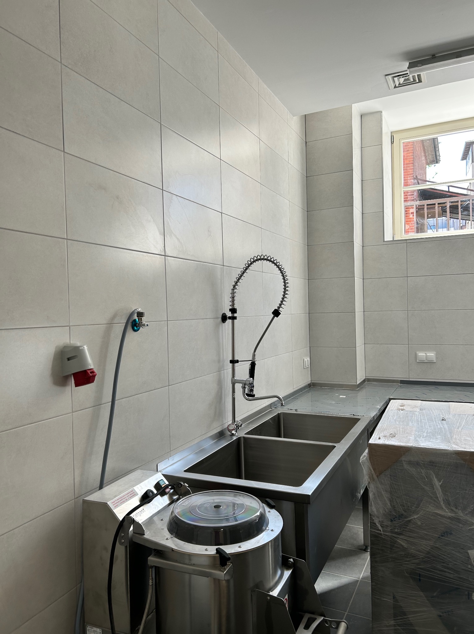 Wnętrze po remoncie: błyszcząca, dwukomorowa umywalka ze stali nierdzewnej, armatura i maszyna do obierania ziemniaków na tle jasnych, kwadratowych płytek.