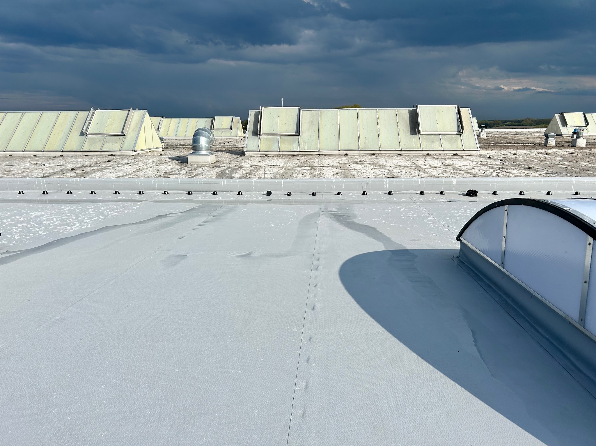 Biały dach przemysłowy z świetlikami i śladami po czyszczeniu, na tle burzowego nieba. Widoczne elementy wentylacji i mocowania. Ujęcie z góry.