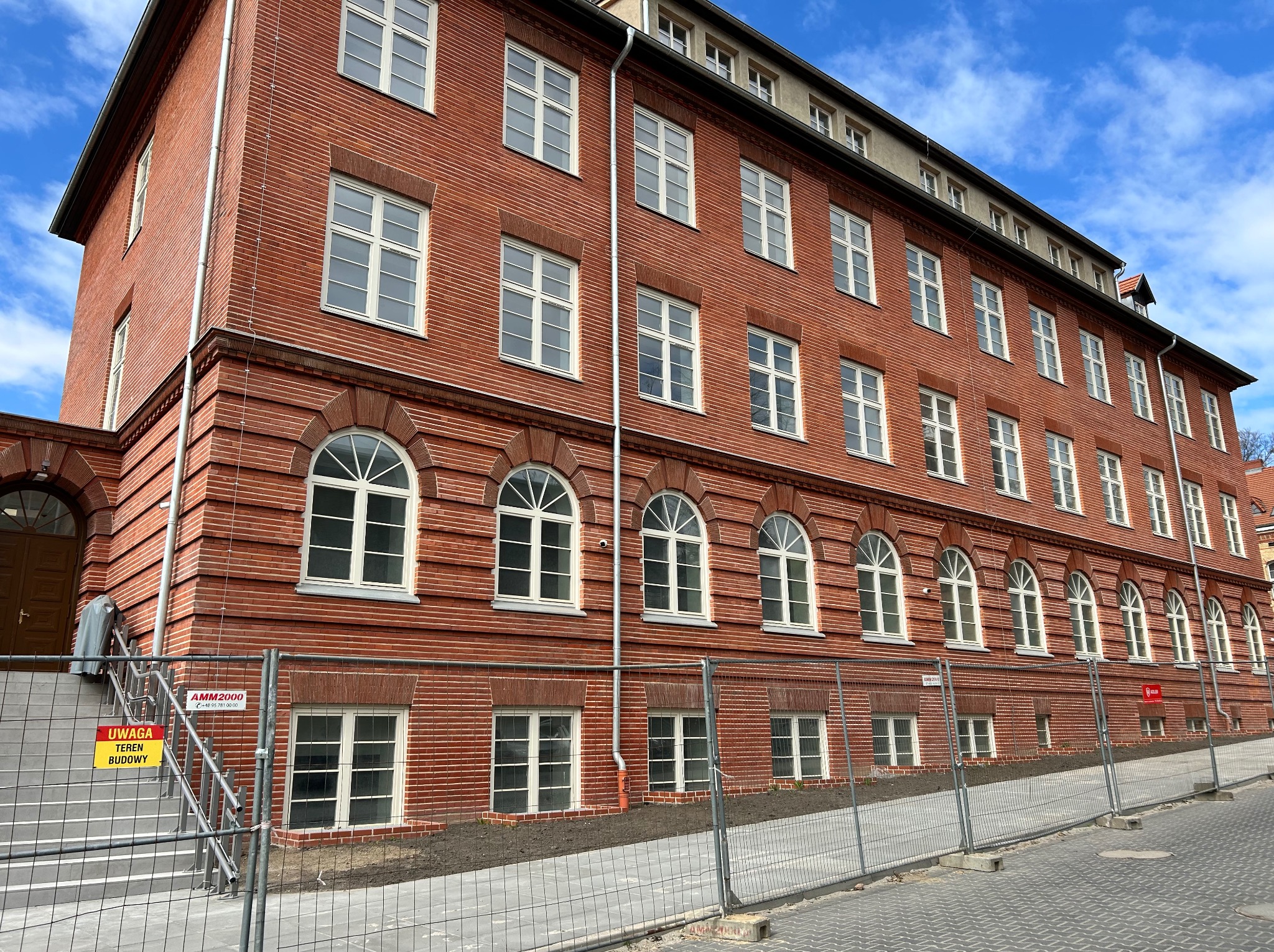Ceglany budynek po renowacji z nowymi oknami, zabezpieczony barierkami budowlanymi i tablicą informacyjną 'Uwaga teren budowy'. Błękitne niebo w tle.