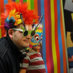 Mężczyzna w okularach w kształcie gitary i kolorowej peruce oraz dziecko w masce na tle kolorowej ścianki i zdjęć z rekwizytami.