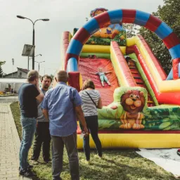 Dziecko z rozłożonymi ramionami zjeżdża z dmuchanej zjeżdżalni w kształcie lwa, obok grupa dorosłych obserwuje zabawę, w tle widoczny teren piknikowy.