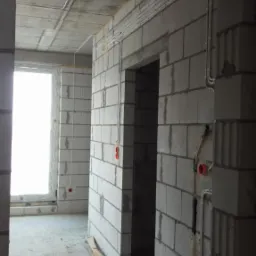 Wnętrze surowego budynku w trakcie prac instalacyjnych: widoczne przewody elektryczne prowadzone po ścianach z bloczków, puszki instalacyjne i betonowy strop.