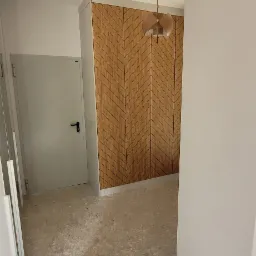 Wnętrze pokoju z drewnianą ścianą w jodełkę, szarą podłogą lastryko i wiszącą lampą z abażurem. Widoczna biała ściana i drzwi.