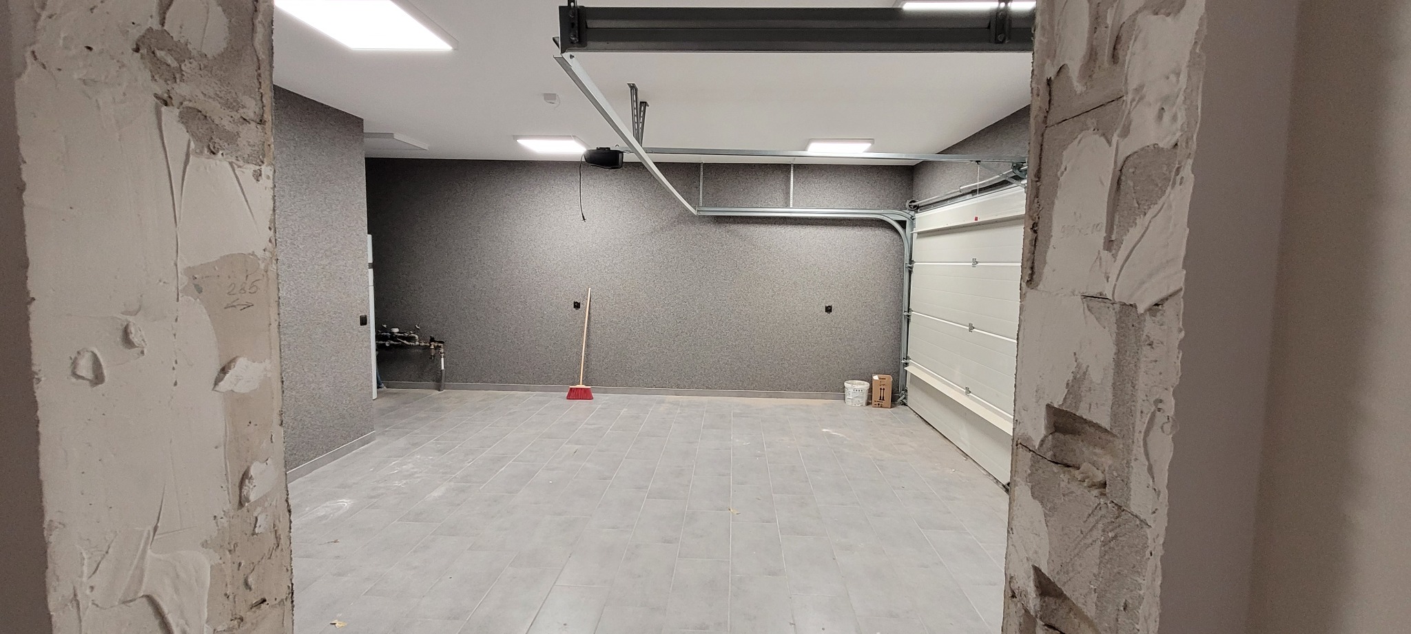Wnętrze garażu w trakcie wykańczania: ściany wykończone tapetą strukturalną, szara płytka podłogowa, brama garażowa częściowo otwarta, widoczne resztki zaprawy na ościeżnicy.