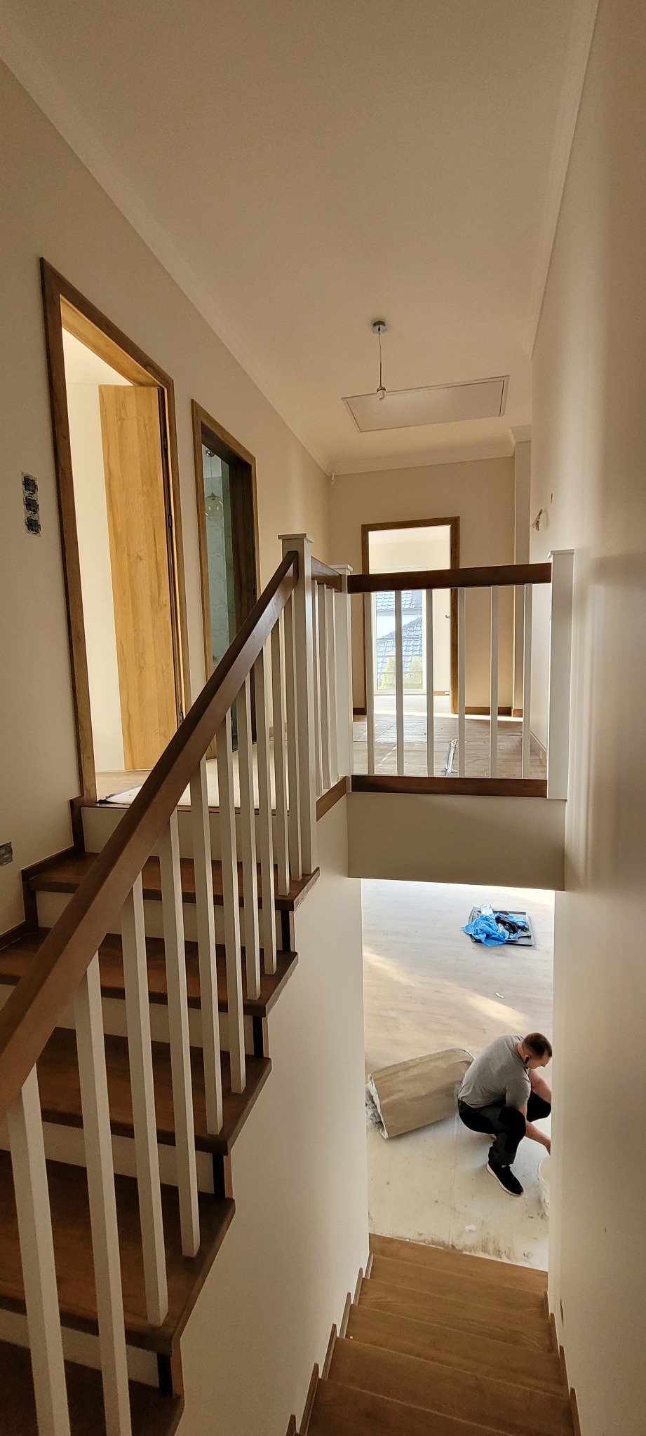 Widok z góry na klatkę schodową z drewnianymi stopniami i białymi balustradami; w tle widoczny pracownik przygotowujący podłoże pod parkiet w jasnym pomieszczeniu.