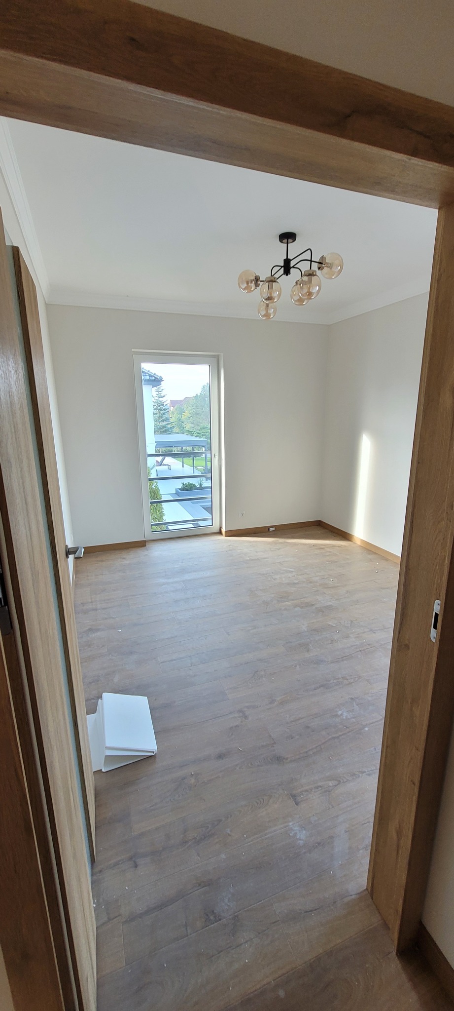 Pusty pokój z drewnianą podłogą, świeżo pomalowanymi białymi ścianami, widokiem na zewnątrz przez okno balkonowe i nowoczesną lampą sufitową, sfotografowany zza drewnianej ościeżnicy drzwiowej.