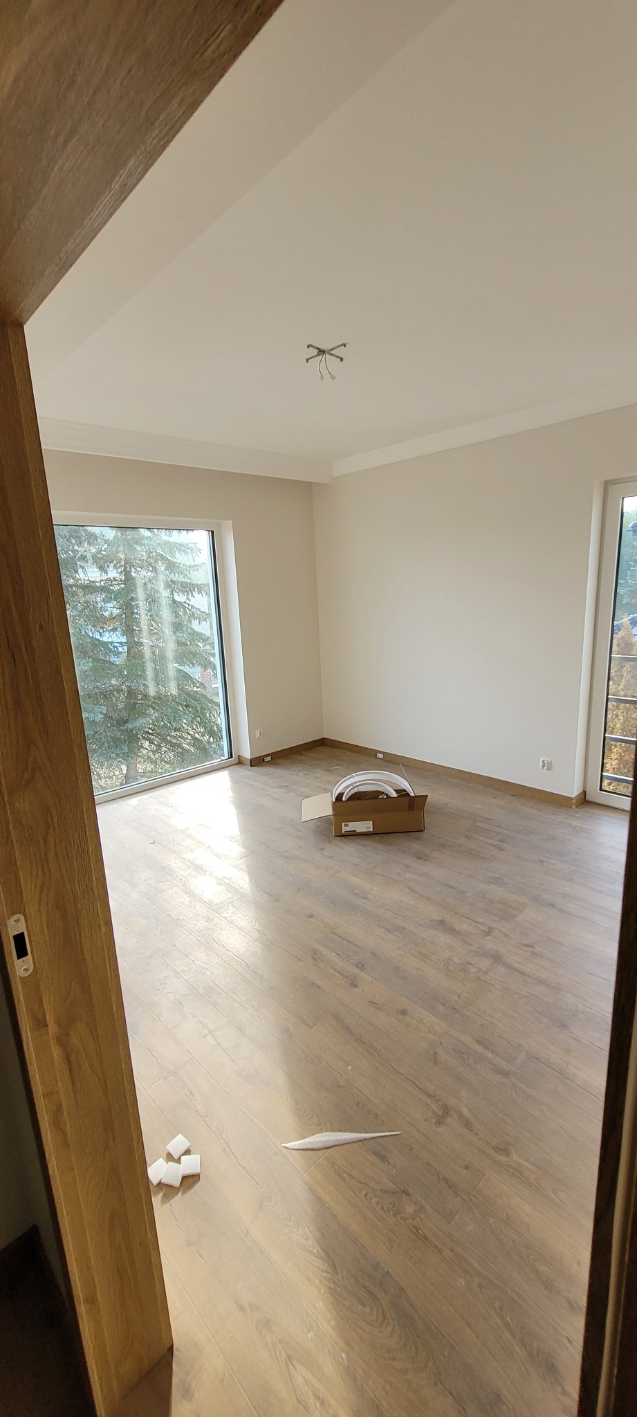 Pusta przestrzeń z panelami podłogowymi, listwami przypodłogowymi, świeżo pomalowanymi ścianami i sufitem oraz kartonem z elementami oświetlenia na podłodze, widok zza futryny drzwi.