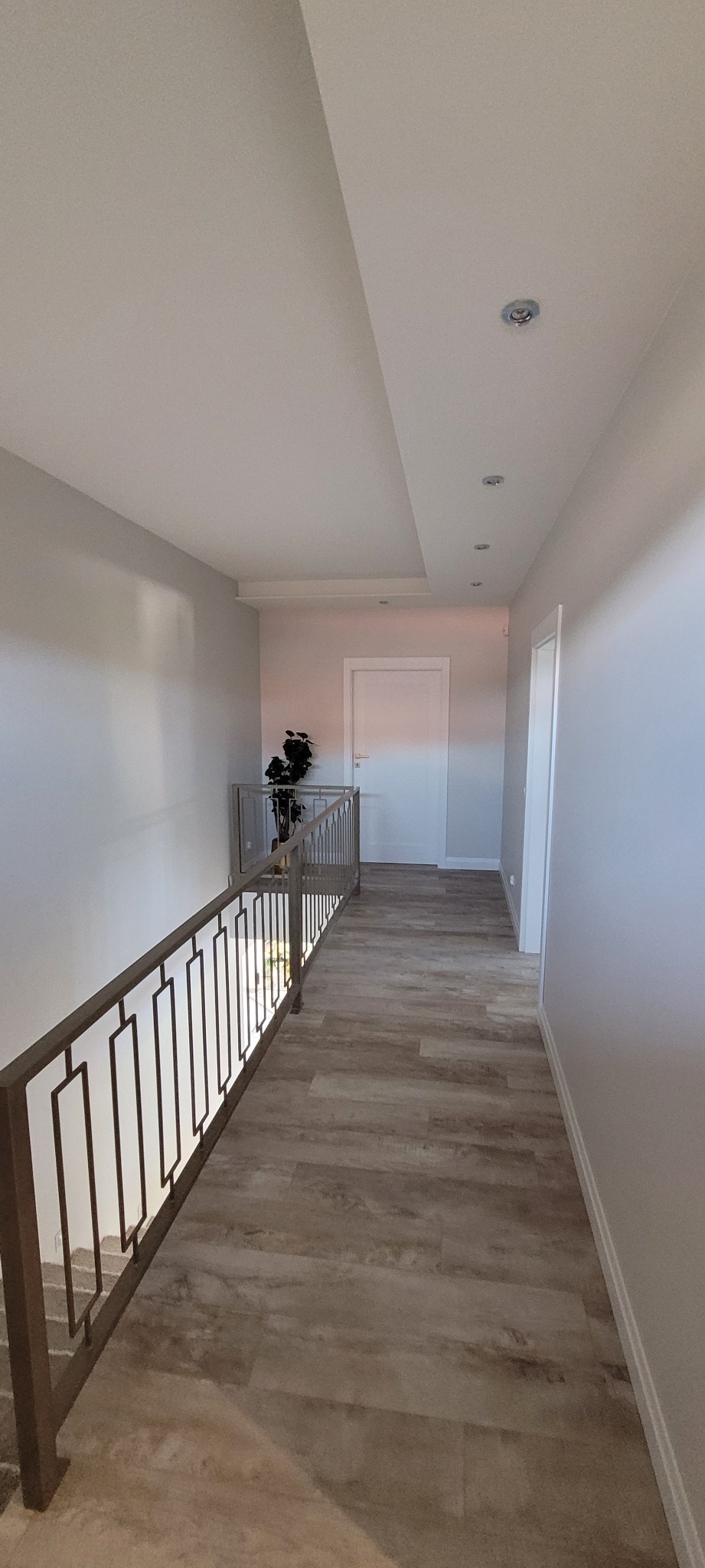 Nowoczesny korytarz na piętrze z drewnopodobną podłogą, metalową balustradą z geometrycznym wzorem i białymi ścianami z listwami przypodłogowymi.