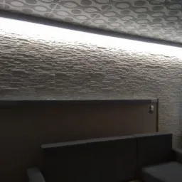 Ściana z dekoracyjnej, strukturalnej okładziny w Pasterzowicach, oświetlona listwą LED pod sufitem, z widocznym fragmentem sufitu z wzorem i narożnikiem sofy na pierwszym planie.