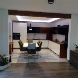 Nowoczesna kuchnia z białymi i czarnymi szafkami, drewnianym blatem i stołem jadalnym z krzesłami, widok z salonu z drewnianą belką stropową.