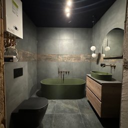 Mieszkanie w Twoim stylu - Nowoczesna łazienka w odcieniach szarości i zieleni z wolnostojącą wanną, umywalką na szafce z drewnianymi lamelami i wiszącą toaletą. Na ścianie grzejnik.