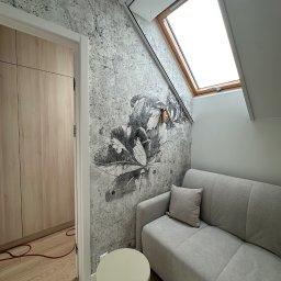 Mieszkanie w Twoim stylu - Pokój na poddaszu z szarą sofą, oknem dachowym i tapetą w kwiaty. Widoczny fragment szafy i stolika. Czerwony kabel na podłodze dodaje koloru.