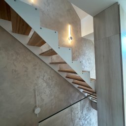 Mieszkanie w Twoim stylu - Nowoczesne schody z drewnianymi stopniami i białymi wspornikami, oświetlone lampami ściennymi, na tle ściany z dekoracyjną fakturą w odcieniach beżu i szarości.