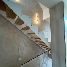 Nowoczesne schody z drewnianymi stopniami i białymi wspornikami, oświetlone lampami ściennymi, na tle ściany z dekoracyjną fakturą w odcieniach beżu i szarości.