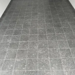 Wykończona podłoga z szarych, kwadratowych płytek gresowych z drobnym wzorem kamyczków, białe fugi, listwa przypodłogowa, widoczne gniazdko elektryczne na ścianie i wejście do innego pomieszczenia.