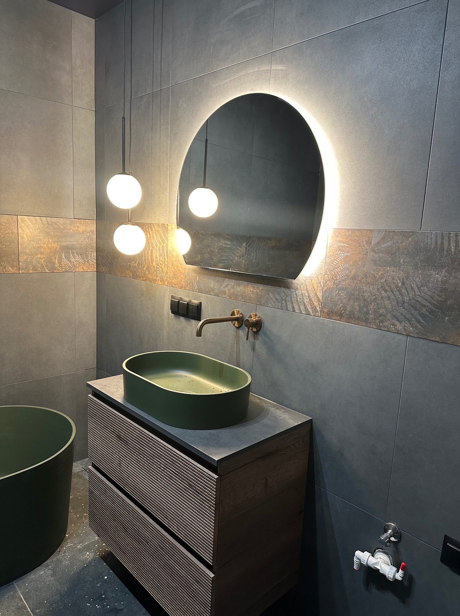 Nowoczesna łazienka z zieloną umywalką nablatową, szafką z lamelami i okrągłym lustrem z podświetleniem. Szare płytki i wiszące lampy kulki.