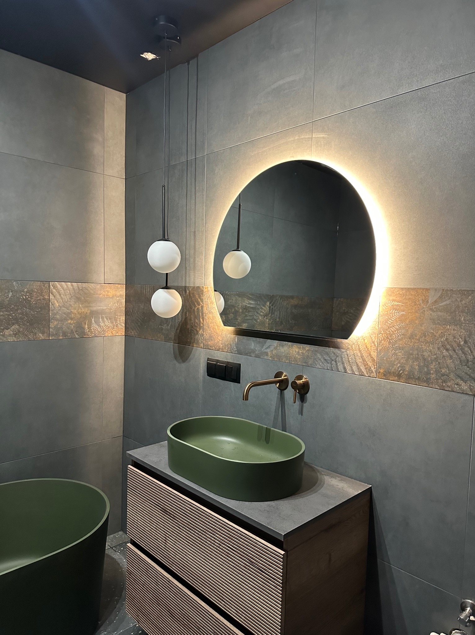 Nowoczesna łazienka z zieloną umywalką nablatową, podświetlanym lustrem i szarą glazurą z dekoracyjnym pasem. Minimalistyczne lampy wiszące.