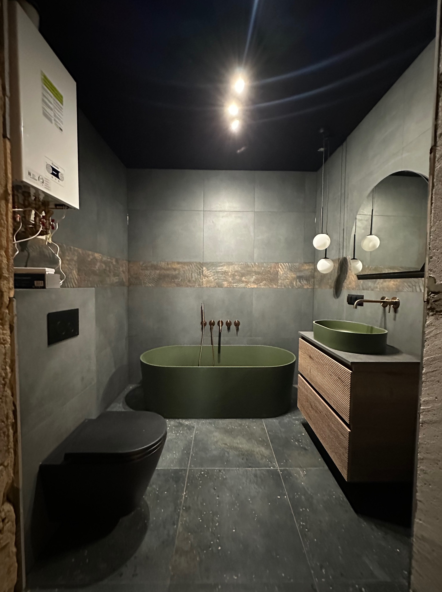 Nowoczesna łazienka w odcieniach szarości i zieleni z wolnostojącą wanną, umywalką na szafce z drewnianymi lamelami i wiszącą toaletą. Na ścianie grzejnik.