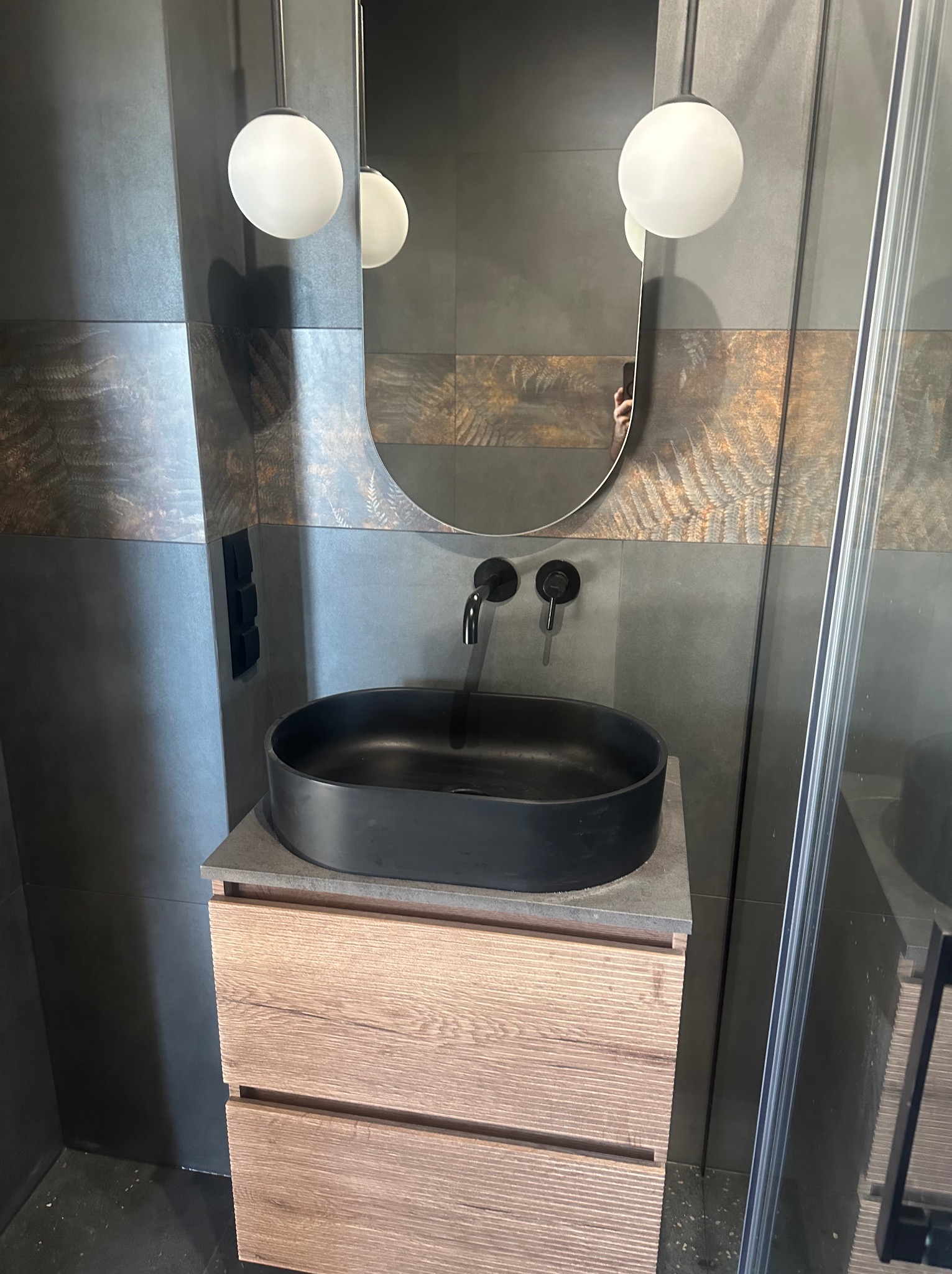 Nowoczesna łazienka: czarna umywalka na drewnianej szafce, szara ściana z roślinnym motywem, okrągłe lampy i owalne lustro. Minimalistyczny design.