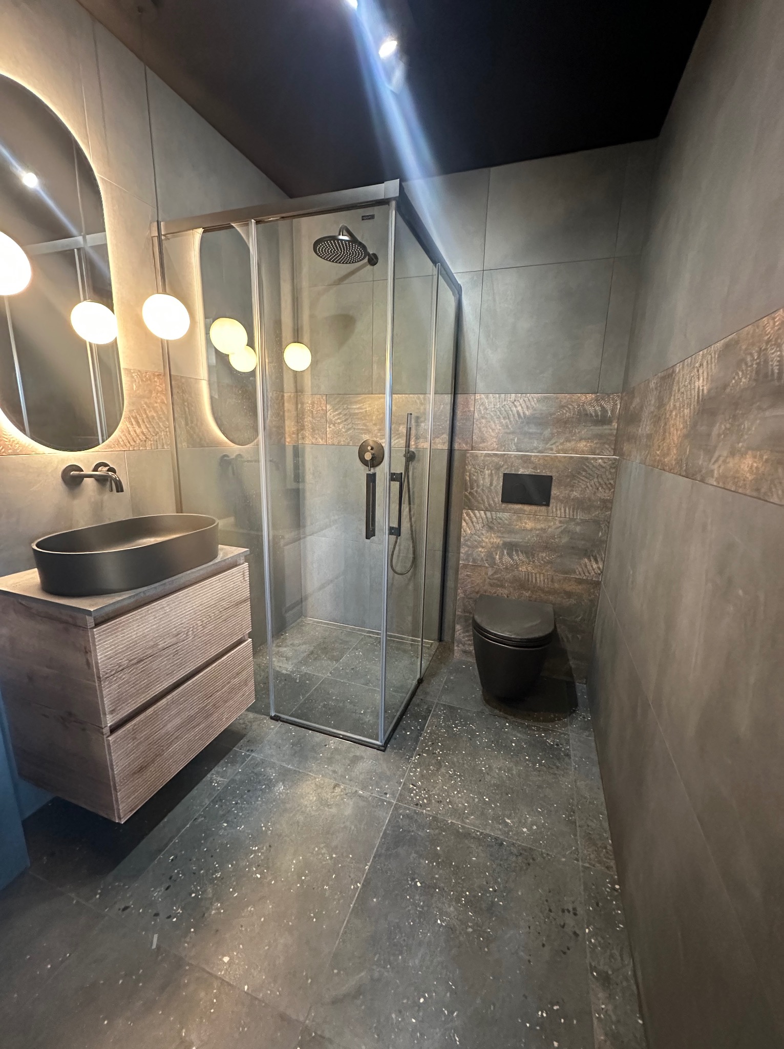 Nowoczesna łazienka w szarościach z kabiną prysznicową, umywalką na szafce z drewnianymi frontami i podświetlanym lustrem. Ciemna armatura i płytki z motywem liści.