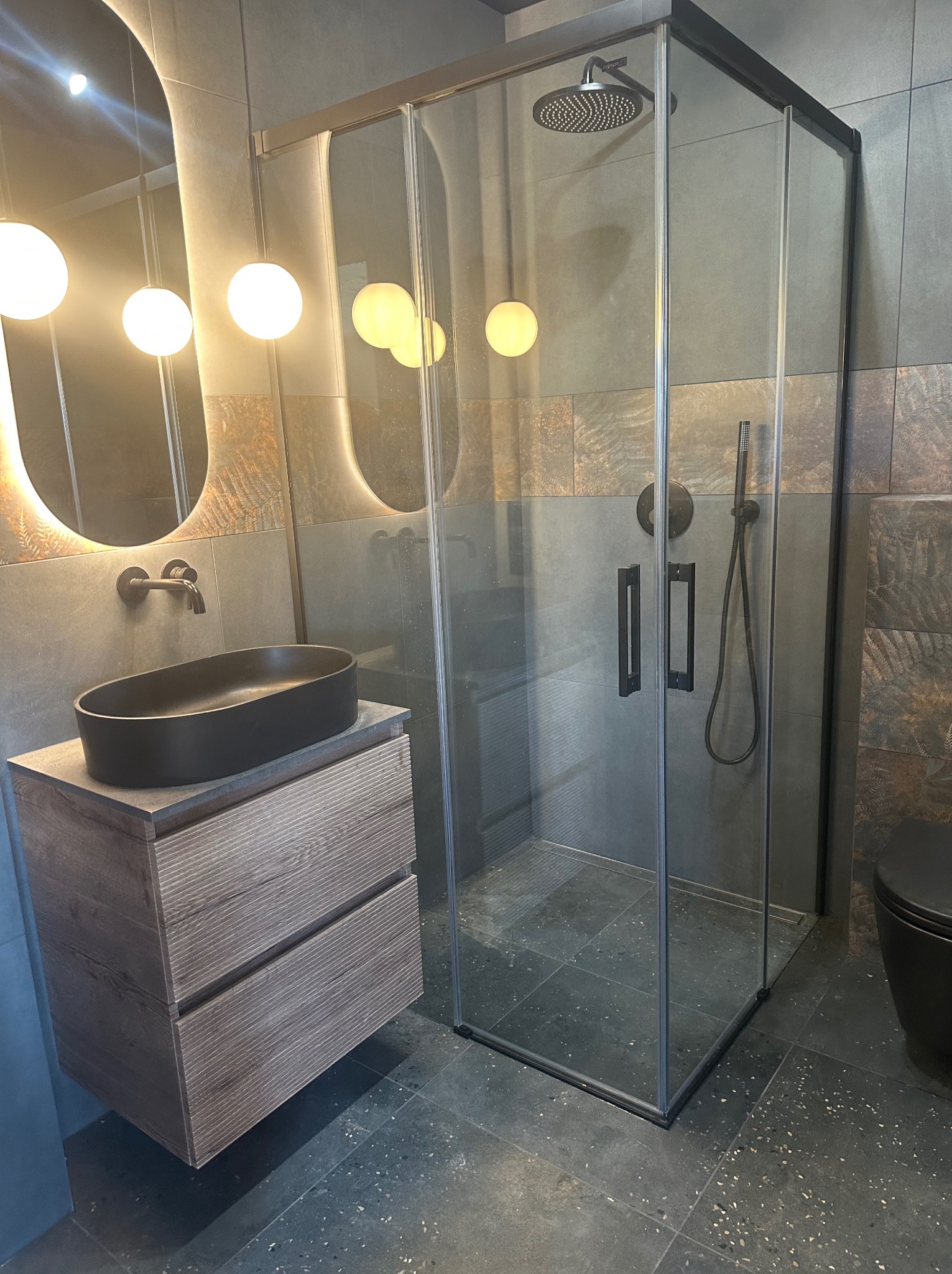 Nowoczesna łazienka w szarościach: kabina prysznicowa z czarnymi ramami, umywalka nablatowa na drewnianej szafce, lustro z podświetleniem i płytki z motywem roślinnym.