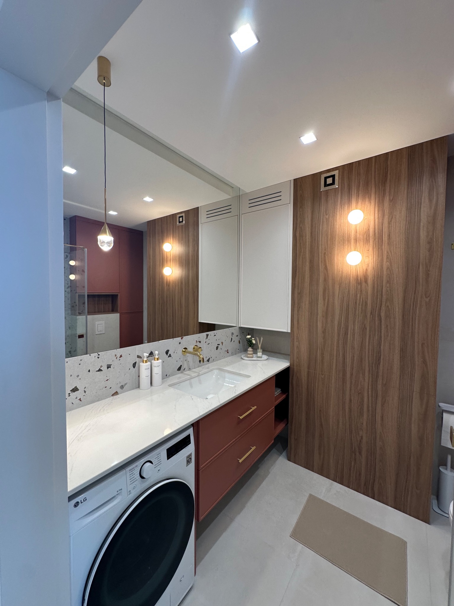 Nowoczesna łazienka z pralką LG, umywalką w blacie z lastryko i szafkami w kolorze terakoty. Na ścianie drewnopodobna okładzina z okrągłymi lampami.