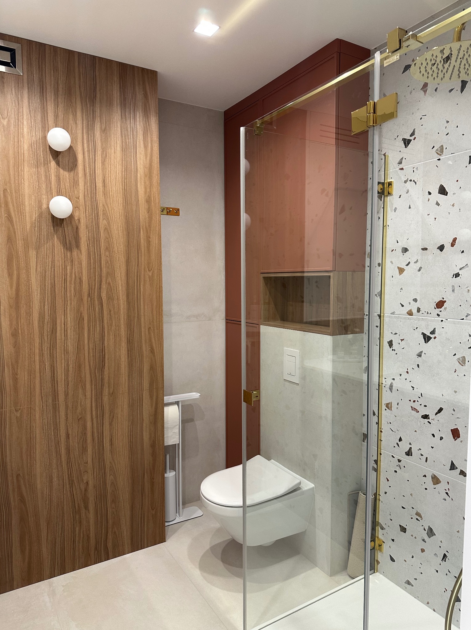 Nowoczesna łazienka z kabiną prysznicową typu walk-in, płytkami lastryko i elementami drewna. Uwagę zwracają złote detale i minimalistyczna toaleta podwieszana.