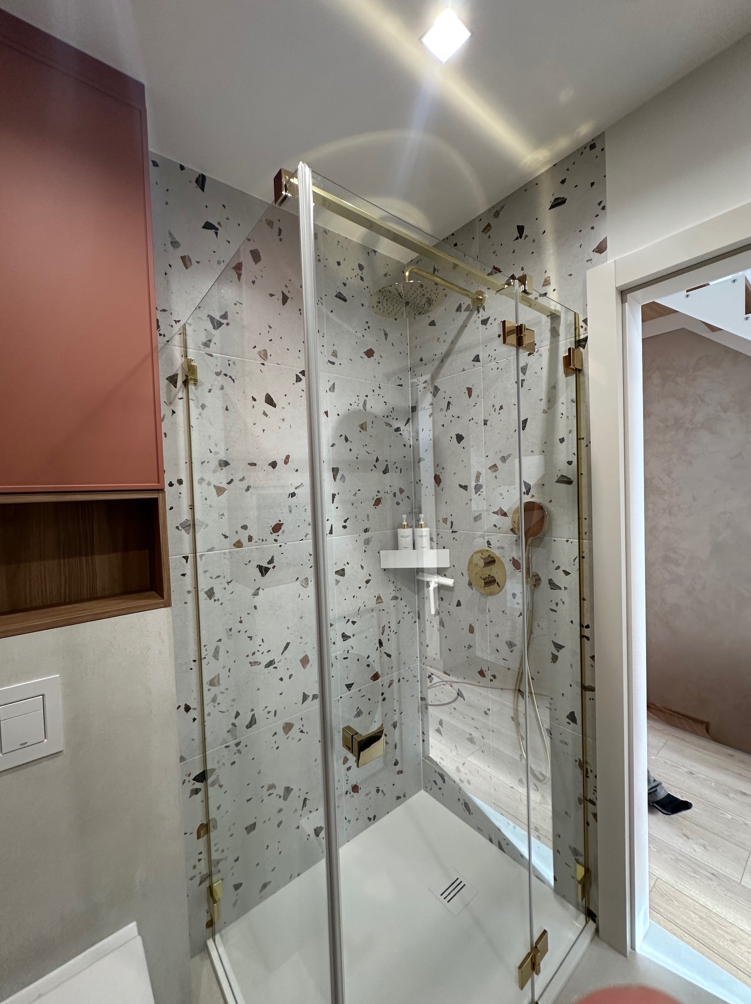 Nowoczesna kabina prysznicowa z złotymi okuciami i mozaiką na ścianach. Widoczny fragment szafki w kolorze terakoty i podłoga wykończona panelami.