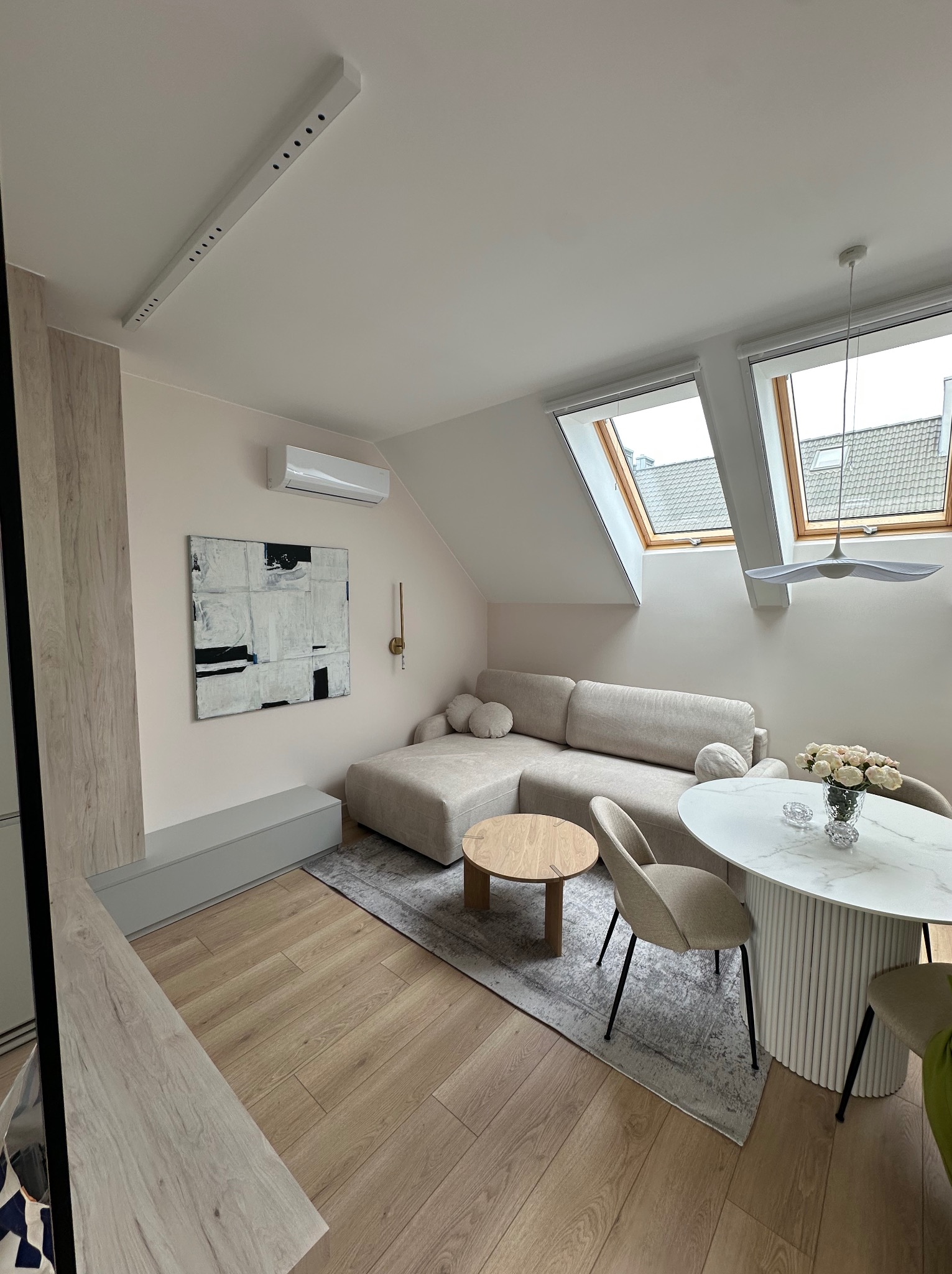 Jasne wnętrze poddasza ze skośnymi oknami: beżowa sofa narożna, okrągły stół z kwiatami, abstrakcyjny obraz na ścianie. Nowoczesny design, ciepłe oświetlenie.