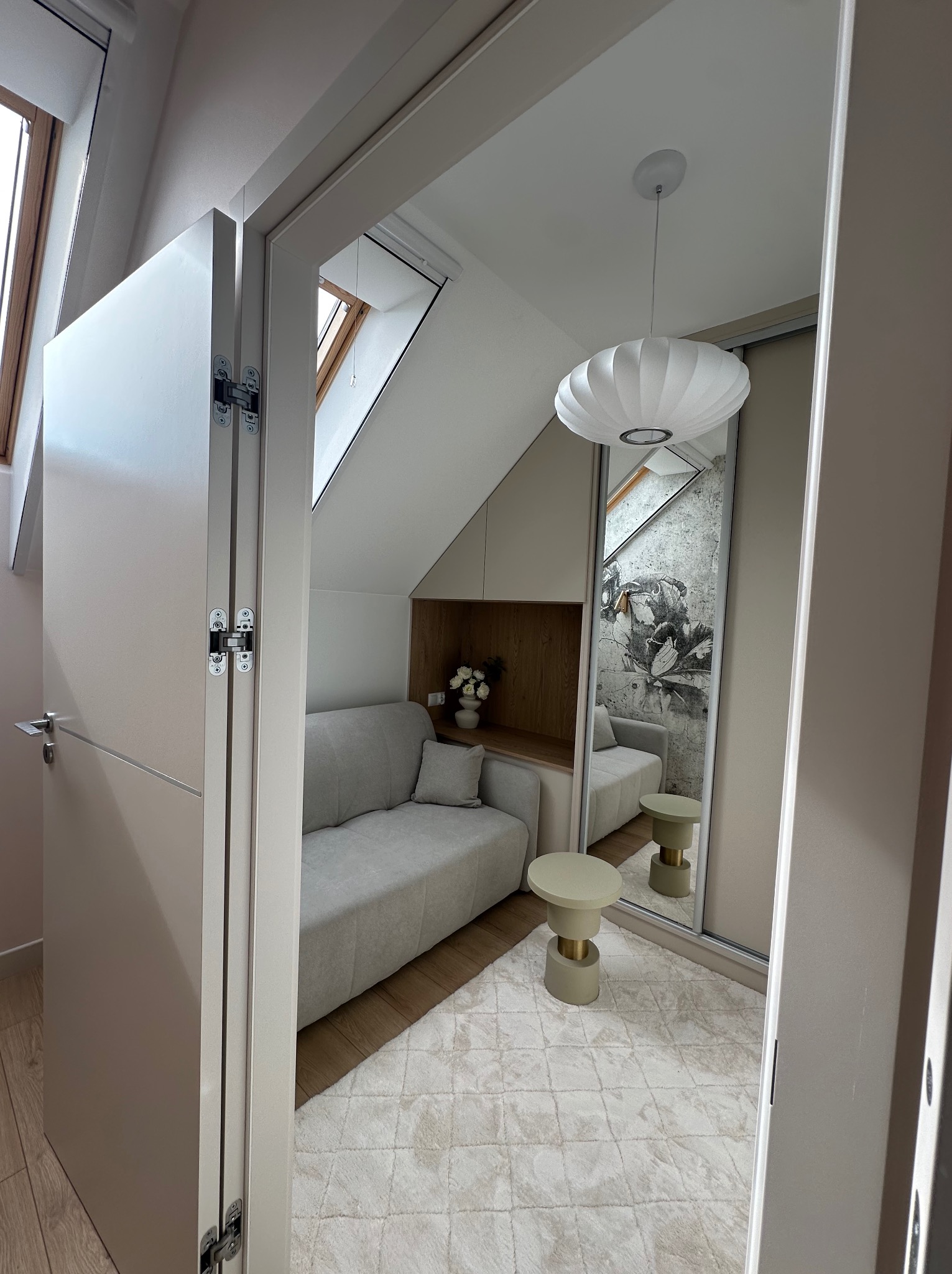 Kompaktowa aranżacja pod skosem: sofa, stolik i lustro powiększające przestrzeń w jasnym wnętrzu z akcentem drewna i dekoracyjną tapetą.