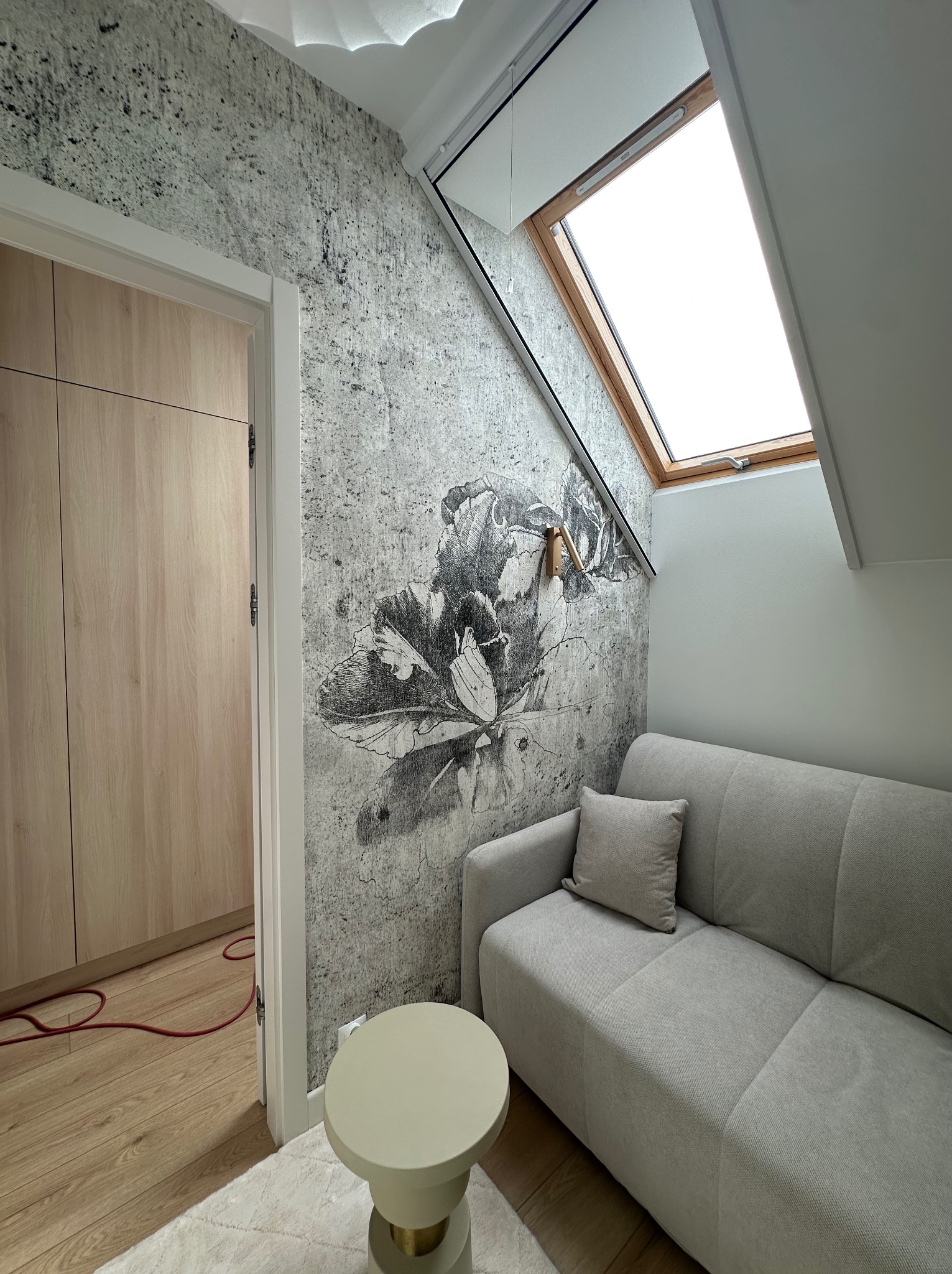 Pokój na poddaszu z szarą sofą, oknem dachowym i tapetą w kwiaty. Widoczny fragment szafy i stolika. Czerwony kabel na podłodze dodaje koloru.
