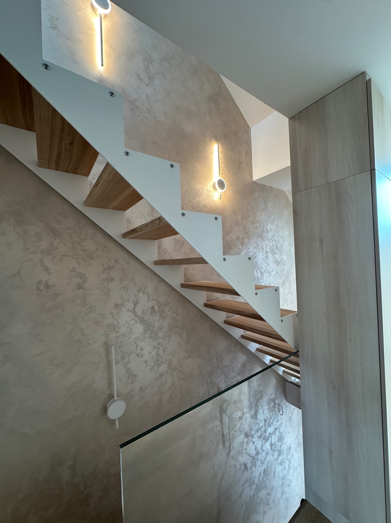Nowoczesne schody z drewnianymi stopniami i białymi wspornikami, oświetlone lampami ściennymi, na tle ściany z dekoracyjną fakturą w odcieniach beżu i szarości.