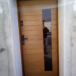 Nowoczesne drzwi wejściowe z poziomymi panelami drewnianymi i pionowym, czarnym panelem szklanym, zamontowane w surowym otworze ściennym z widocznymi śladami zaprawy.